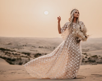May include: A woman wearing a white lace gown with a crown of thorns and holding a bouquet of dried flowers stands in a desert landscape.