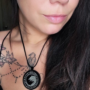 Pu&ograve; includere: Una donna con lunghi capelli neri indossa una camicetta nera con scollo a pizzo. Indossa una collana con un ciondolo rotondo che presenta un design a lupo e rune.