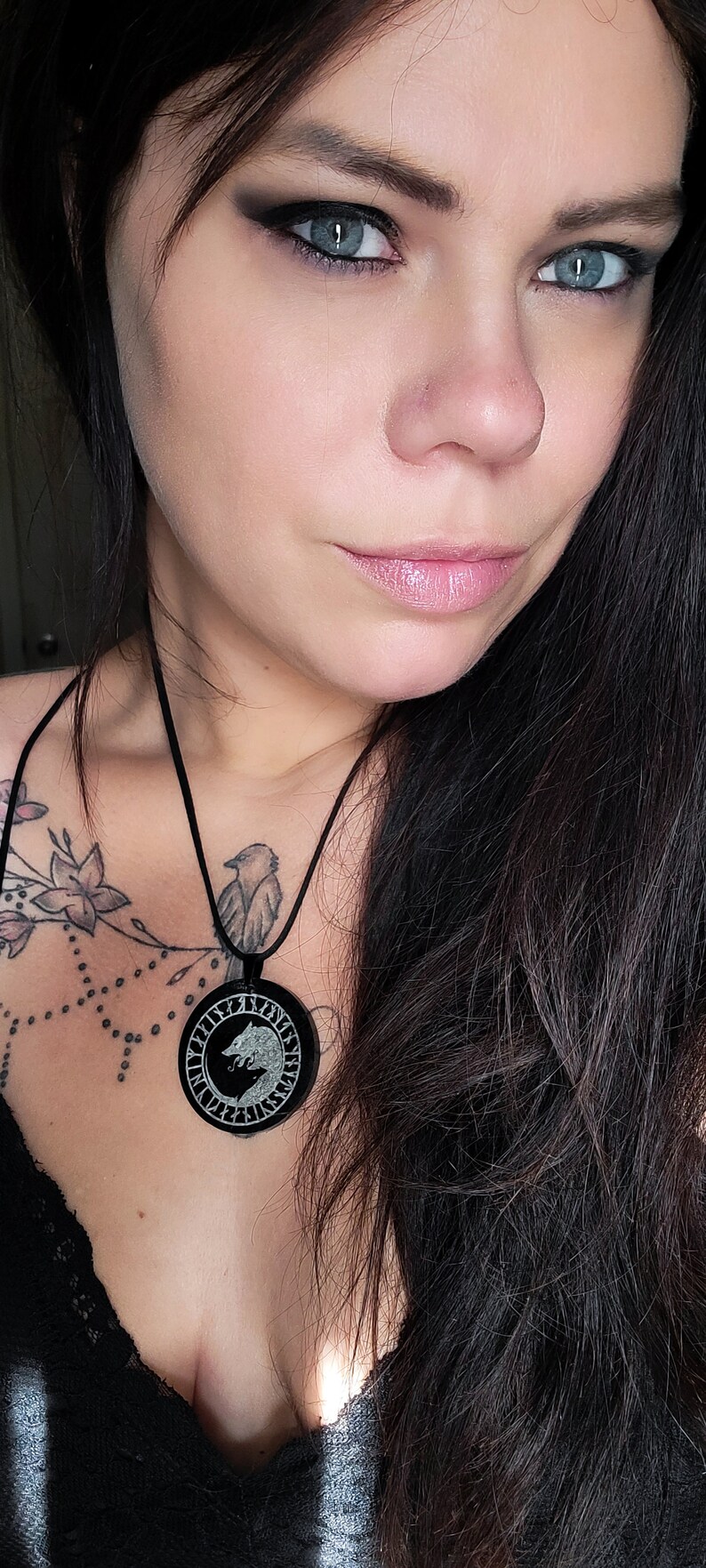 Pu&ograve; includere: Una donna con lunghi capelli neri indossa una camicetta nera con scollo a pizzo. Indossa una collana con un ciondolo rotondo che presenta un design a lupo e rune.