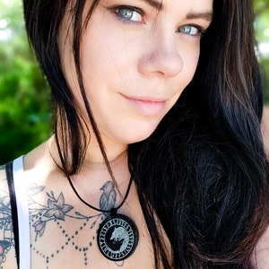 Pu&ograve; includere: Una donna con lunghi capelli neri indossa una canotta bianca con un tatuaggio nero sul petto. Indossa una collana con un ciondolo rotondo che presenta un design a lupo e rune.