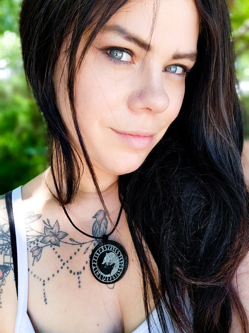 Pu&ograve; includere: Una donna con lunghi capelli neri indossa una canotta bianca con un tatuaggio nero sul petto. Indossa una collana con un ciondolo rotondo che presenta un design a lupo e rune.