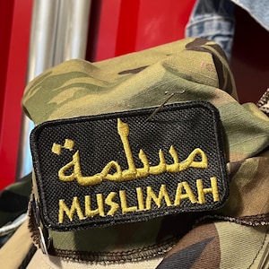 May include: A black patch with the word "MUSLIMAH" embroidered in gold, with Arabic script above it. The patch is pinned to a camouflage fabric, with a tan and green hat visible below.
