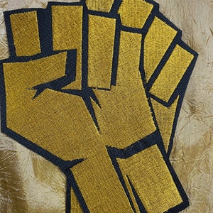 May include: Embroidered patch featuring a raised fist design. The fist is rendered in gold thread with a black outline. The patch is likely intended for application on clothing or accessories, symbolizing solidarity or activism.