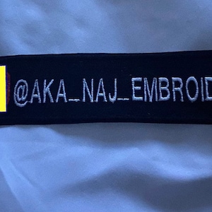 Op de afbeelding: Een zwarte geborduurde patch met witte stiksels met de tekst "@AKA_NAJ_EMBROIDERY".