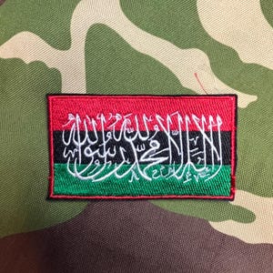 Peut inclure: Écusson brodé avec un motif rouge, noir et vert. L'écusson présente une écriture arabe blanche et une bordure noire. Le fond est un motif de camouflage.