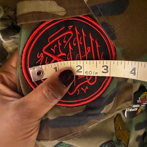 May include: A black and red embroidered patch with Arabic script in a circle. The patch is attached to a camouflage fabric. A measuring tape is used to measure the patch, which is approximately 2 inches in diameter.