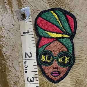 May include: Embroidered patch featuring a black woman with a red, green, and yellow head wrap, wearing large round sunglasses with the word "BLACK" written across the lenses.