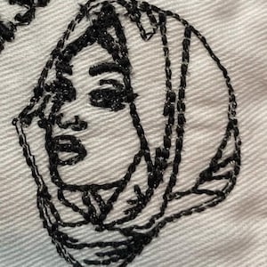 May include: Close-up of a white fabric with a black embroidered face design. The embroidery depicts a woman's face with detailed features, including eyes, nose, and lips. The design is stitched with thick, dark thread, creating a bold outline.