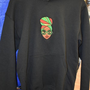May include: Black hooded sweatshirt with a front pocket and drawstring hood. The sweatshirt features an embroidered design of a woman's face wearing sunglasses with the word "BLACK" on the lenses, and a colorful head wrap.