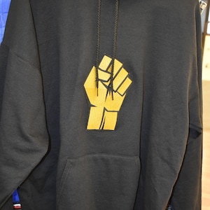 May include: Black hooded sweatshirt with a gold raised fist graphic on the front. The hoodie has a drawstring and a kangaroo pocket.