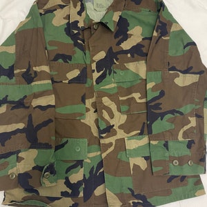 May include: A green, brown, and black camouflage military jacket with a button-up front and two large pockets on the front.