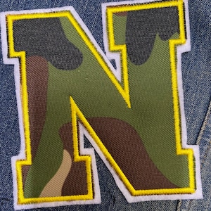 May include: A large, white felt letter N with a yellow outline. The letter is filled with a camouflage pattern in shades of green, brown, and black.