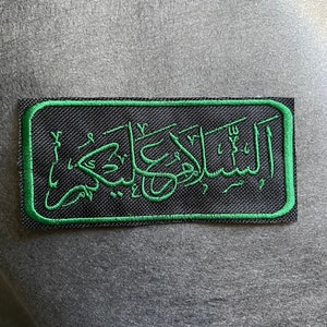 May include: A black patch with a green embroidered border. The patch features Arabic calligraphy in green thread.