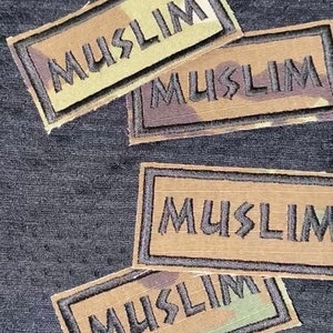 Könnte beinhalten: Vier braune und schwarze gestickte Patches mit dem Wort "MUSLIM" in schwarzen Buchstaben auf einem Tarnmuster-Hintergrund.