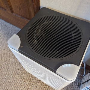 May include: A white air purifier with a black top and a circular air intake grill. The air purifier has a small white light on the top right corner.
