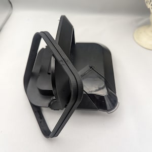 Puede incluir: Un dispositivo de plástico negro con una correa de goma curvada. El dispositivo tiene una base rectangular y un componente de plástico transparente. La correa está enrollada alrededor del dispositivo.