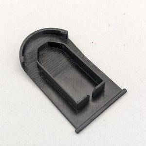 Puede incluir: Un cortador de galletas negro con forma de ataúd, impreso en 3D. El cortador tiene una base rectangular con una parte superior redondeada y un contorno de ataúd en el centro. El diseño es de plástico negro.