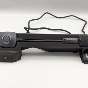 May include: A black plastic headset with a microphone and a control button. The headset has a black cord and a black strap.