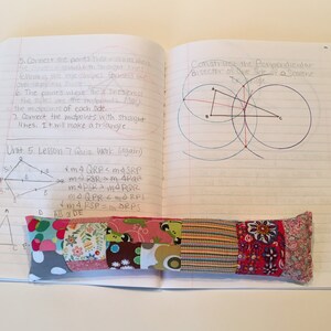 May include: A notebook with handwritten notes on geometry, including diagrams and equations. The notebook is open to a page titled "Unit 5. Lesson 7 Quiz Work (Again)". A colorful fabric pencil holder is resting on the notebook.