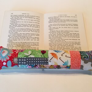 May include: A multi-colored fabric book mark with a variety of patterns, including flowers, umbrellas, and geometric shapes. The bookmark is resting on an open book.