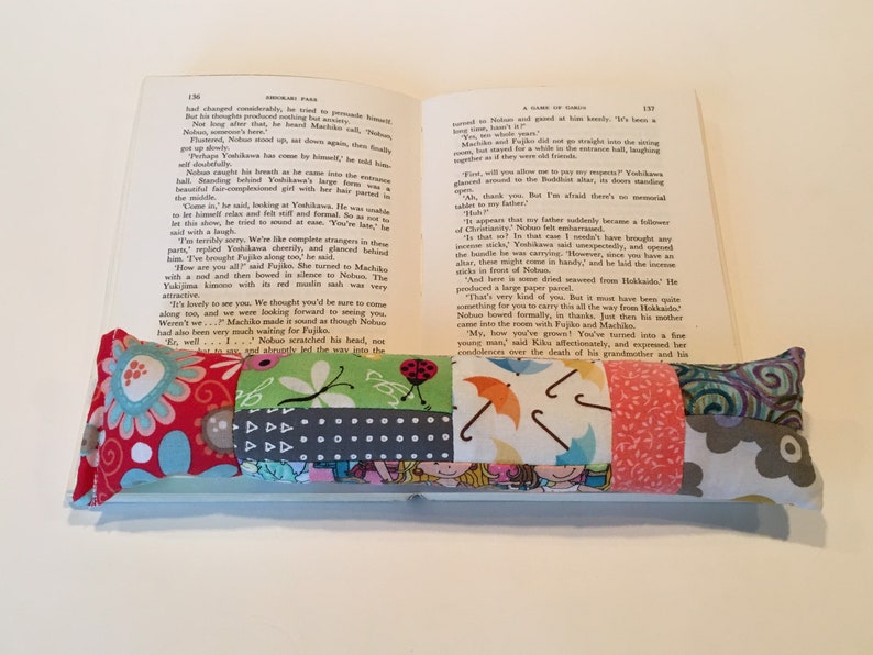 May include: A multi-colored fabric book mark with a variety of patterns, including flowers, umbrellas, and geometric shapes. The bookmark is resting on an open book.