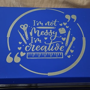 May include: A blue storage box with a silver design that reads "I'm not messy, I'm creative." The design includes scissors, a pen, a ruler, and hearts. The box is rectangular and appears to be made of metal.