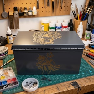 May include: A rectangular dark gray metal storage box featuring a gold-colored graphic of a character in a skirt on the front and a floral design on the lid. The box has a hinged lid with a visible metal hinge running along the back edge.