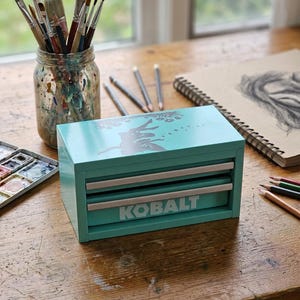 May include: A turquoise metal storage chest with two drawers and the word KOBALT printed in white on the bottom drawer. The top surface features a dark silhouette graphic of a fairy surrounded by small flowers and stars in a light teal color.
