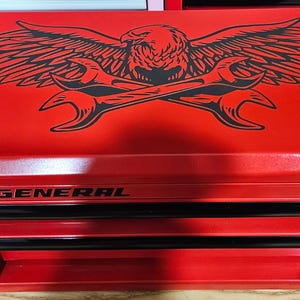 May include: A red metal tool chest with three drawers. The top of the chest features a black graphic of an eagle with outstretched wings and crossed wrenches. The text "U.S. GENERAL" is printed on the front.