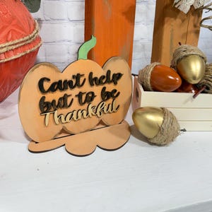 Puede incluir: Un letrero de madera con forma de calabaza con el texto "Can't help but to be Thankful". La imagen también presenta calabazas decorativas de madera, bellotas y cuerda, todo en colores otoñales. Una caja de madera blanca contiene las bellotas.