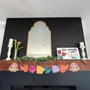 May include: A wooden mantelpiece with a black background. A gold-framed arched mirror hangs above a garland of green leaves. A sign with the text "Give yourself some Grace Today" is displayed on the right side of the mantel. A colorful banner with the word "SPRING" is strung across the front of the mantel.