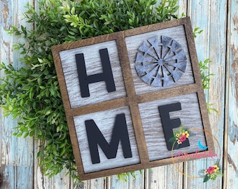 May include: A wooden sign with the word "HOME" spelled out in black letters on a whitewashed background. The sign is divided into four squares, with a metal windmill design in the top right square.