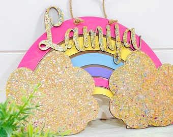 Könnte beinhalten: Ein regenbogenförmiges Holzschild mit dem Namen "Lamiya" in goldenen Glitzerbuchstaben. Der Regenbogen ist in Schichten aus rosa, gelber, blauer und violetter Farbe gehalten. Zwei Wolken mit goldenem Glitzer befinden sich auf beiden Seiten des Regenbogens.