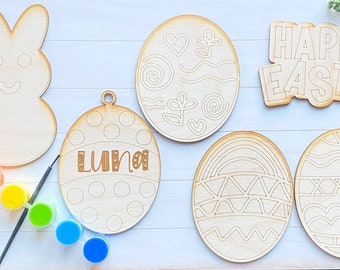 May include: A set of six unfinished wooden Easter craft ornaments. The ornaments include a bunny, an egg with polka dots, an egg with a rainbow design, an egg with a geometric design, an egg with swirls and a heart, and a sign that says "Happy Easter". The ornaments are ready to be painted.