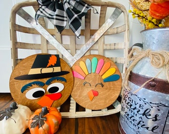 Può includere: Decorazioni in legno a tema Thanksgiving: un tacchino colorato e un pellegrino. Il tacchino ha un ventaglio di piume colorate, mentre il pellegrino indossa un cappello nero. Un cesto decorativo con un fiocco a scacchi e zucche completano l'arredamento autunnale.