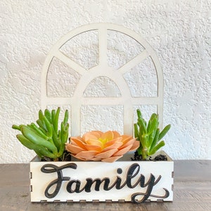 May include: A white wooden planter box with the word "Family" in black script, holding three succulents. The planter is set against a white arched window-shaped backdrop. The succulents are green and peach.