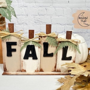 May include: Wooden FALL sign with pumpkin-shaped letters. Each letter is cream-coloured with black lettering, topped with a brown stem, raffia bow, and greenery. A white pumpkin and autumn leaves are in the background.