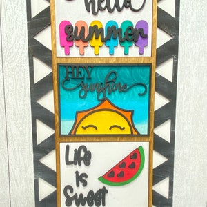 Könnte beinhalten: Ein Holzschild mit drei Tafeln, die sommerliche Texte und Bilder zeigen. Die obere Tafel sagt "hello summer" mit bunten Eis am Stiel-Bildern. Die mittlere Tafel sagt "hey sunshine" mit einer gelben Sonne und blauem Himmel. Die untere Tafel sagt "life is sweet" mit einer roten Wassermelonen-Scheibe.