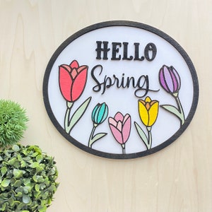 Puede incluir: Un letrero redondo de madera con marco negro y el texto "Hello Spring" en letras negras. El letrero está decorado con cinco tulipanes de colores.