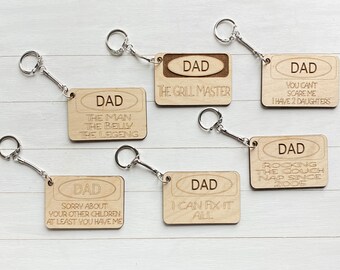 Op de afbeelding: Zes houten sleutelhangers met het woord "DAD" en verschillende humoristische spreuken. De spreuken zijn: "The Grill Master", "The Man The Belly The Legend", "You Can't Scare Me I Have 2 Daughters", "Rocking The Couch Nap Since 2005", "Sorry About Your Other Children At Least You Have Me" en "I Can Fix It All".