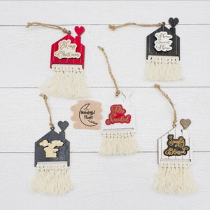 Pode incluir: Cinco enfeites de Natal de madeira em forma de casas com franjas de borla. Os enfeites são pintados de branco e preto com letras douradas. Os enfeites dizem "Merry Christmas", "Home Sweet Home", "Feliz Navidad", "Simply Blessed", e "Moonlight Studio".
