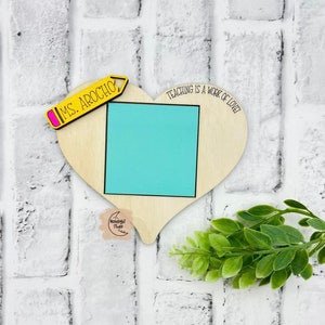 May include: Wooden heart-shaped sign with a light blue square in the center. The sign says "Teaching is a work of love!" and has a yellow pencil with the text "Ms. Arocho" on it.