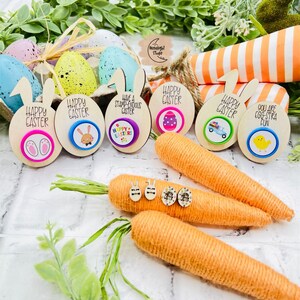 May include: A set of eight wooden Easter egg decorations with bunny ears and colourful buttons. The decorations feature various Easter-themed designs, including bunnies, eggs, and a truck. The decorations are displayed on a background of carrots and green foliage.