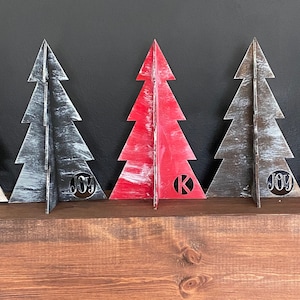 May include: Five wooden Christmas tree decorations in various colors, including white, black, red, brown, and pink. Each tree has a circle cutout in the center with the word "JOY" or a letter.