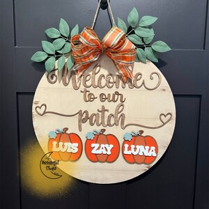 May include: A round wooden sign with the text "Welcome to our patch" and three pumpkins with the names "Luis", "Zay", and "Luna" on them. The sign is decorated with a plaid bow and green leaves.