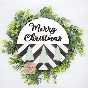 Pode incluir: Um sinal redondo de madeira com um padrão de chevron preto e branco e três árvores verdes. O sinal diz "Merry Christmas" em letras cursivas pretas.