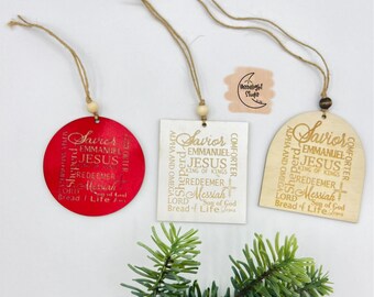 May include: Three wooden Christmas ornaments with a gold inscription of "Savior" and other religious words. The ornaments are in the shapes of a circle, a square, and an arch. The circle ornament is red, the square ornament is white, and the arch ornament is natural wood.
