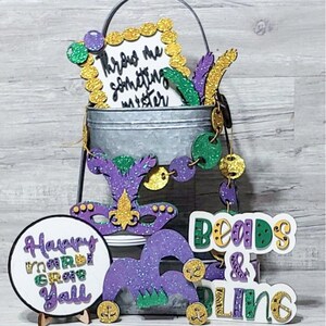 May include: A Mardi Gras themed party decoration set with a purple, green, and gold color scheme. The set includes a sign that says "Throw me something mister", a sign that says "Happy Mardi Gras Y'all", a sign that says "Beads & Dline", a purple and green jester hat, and a purple and gold mask.