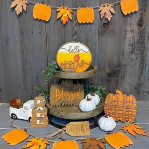 以下が含まれることがあります： 「ハローフォール」、「祝福」、「集まれ」と書かれた看板が置かれた、段になったトレイのある木製秋の装飾。トレイは、白いカボチャ、格子模様のカボチャ、おもちゃのトラックで飾られています。オレンジ色のカボチャと葉がついた秋の横断幕がトレイの上にかけられています。