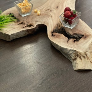 May include: A wooden serving tray with a natural wood grain finish. The tray has a unique shape with a large, irregular centre section. Two small glass bowls are placed on the tray, one containing cubed cheese and the other containing fresh strawberries.